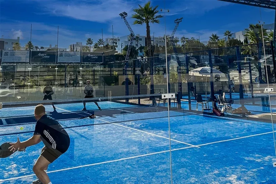 Man hitting a ball on a blue padel court surrounded by palm trees